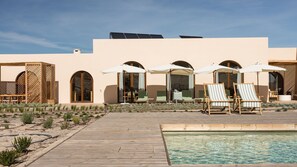 Zona de piscina amb gandules i ombrelles, un pont de fusta i un edifici amb finestres d'arc.