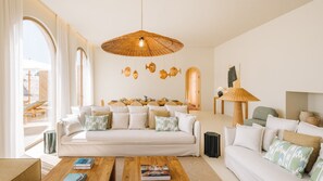 A modern living room with a white sofa, a coffee table, and a dining area with a wooden table and chairs. - Ando Living - Comporta Villas - Villa 4 (Carvalhal)