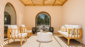 A sunlit patio with wooden furniture, a round table, and a view through an arched window. - Ando Living - Comporta Villas - Villa 7 (Carvalhal)