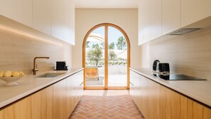A modern kitchen with a central island, a sink, and a view through a large archway to an outdoor area. - Ando Living - Comporta Villas - Villa 11 (Carvalhal)