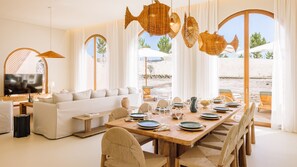 A modern interior with a dining area, sofa, and large windows overlooking a beach. - Ando Living - Comporta Villas - Villa 11 (Carvalhal)
