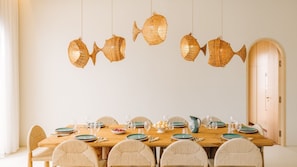 A dining area with a wooden table set for six, surrounded by wicker chairs, with hanging fish-shaped light fixtures above. - Ando Living - Comporta Villas - Villa 17 (Carvalhal)