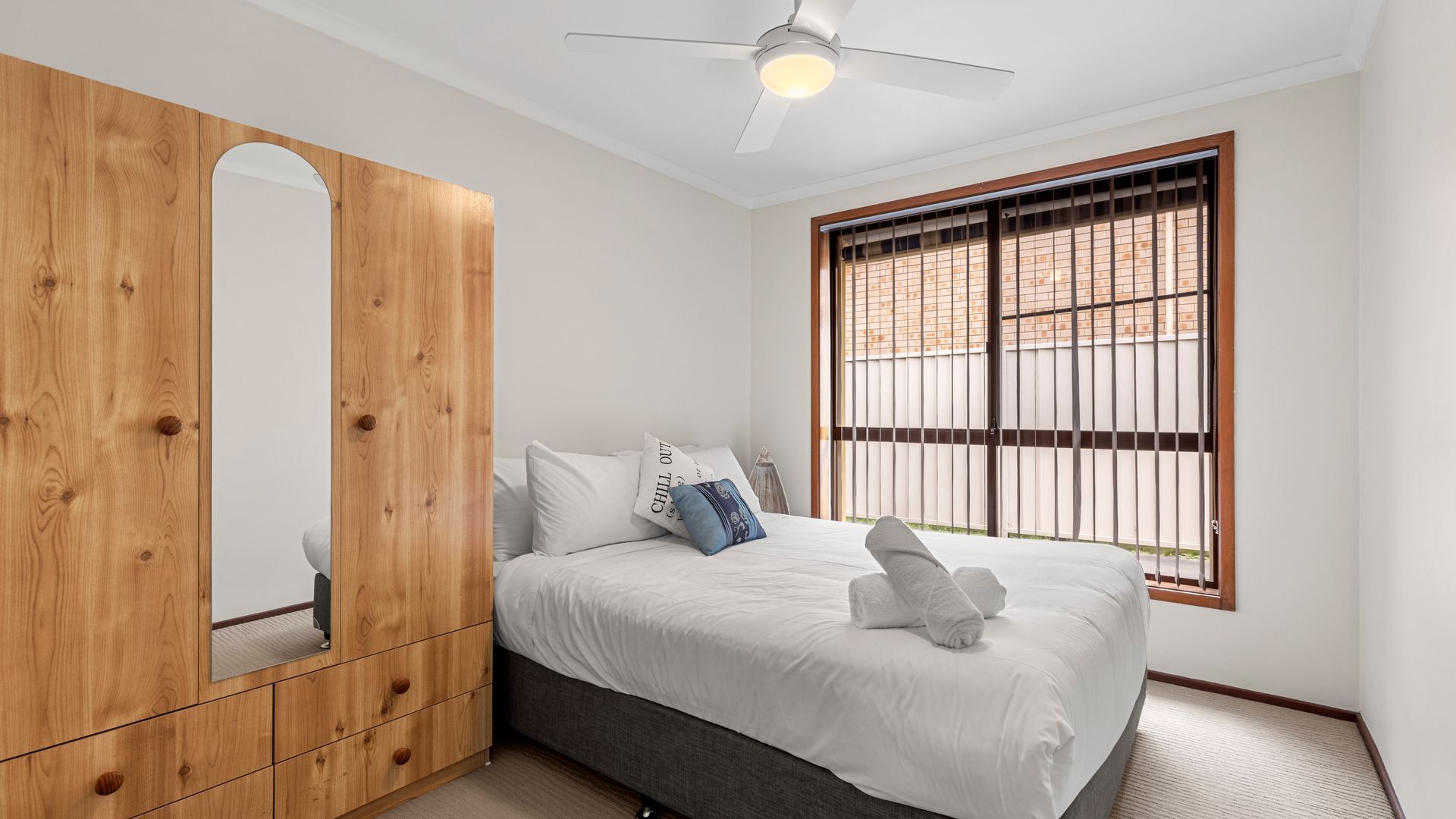 A bedroom with a bed, a wooden wardrobe, a ceiling fan, and a window with metal bars.