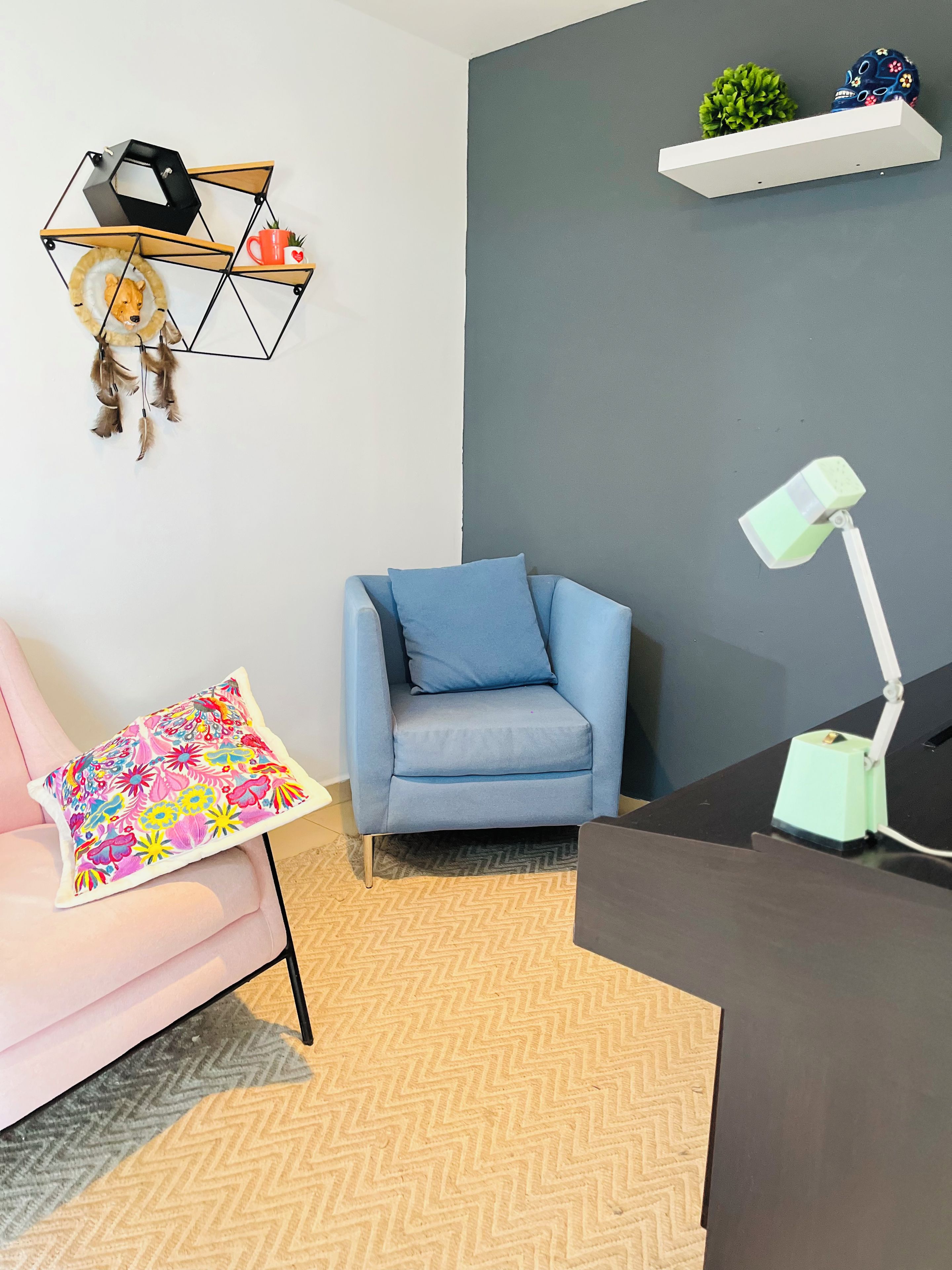 Een kamer met een blauwe fauteuil, een bureau met lamp, een gestreept kussen, een geometrisch vloerkleed en een wandplank met decoratieve objecten.