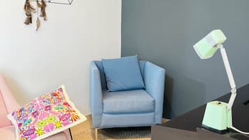 Een kamer met een blauwe fauteuil, een bureau met lamp, een gestreept kussen, een geometrisch vloerkleed en een wandplank met decoratieve objecten.