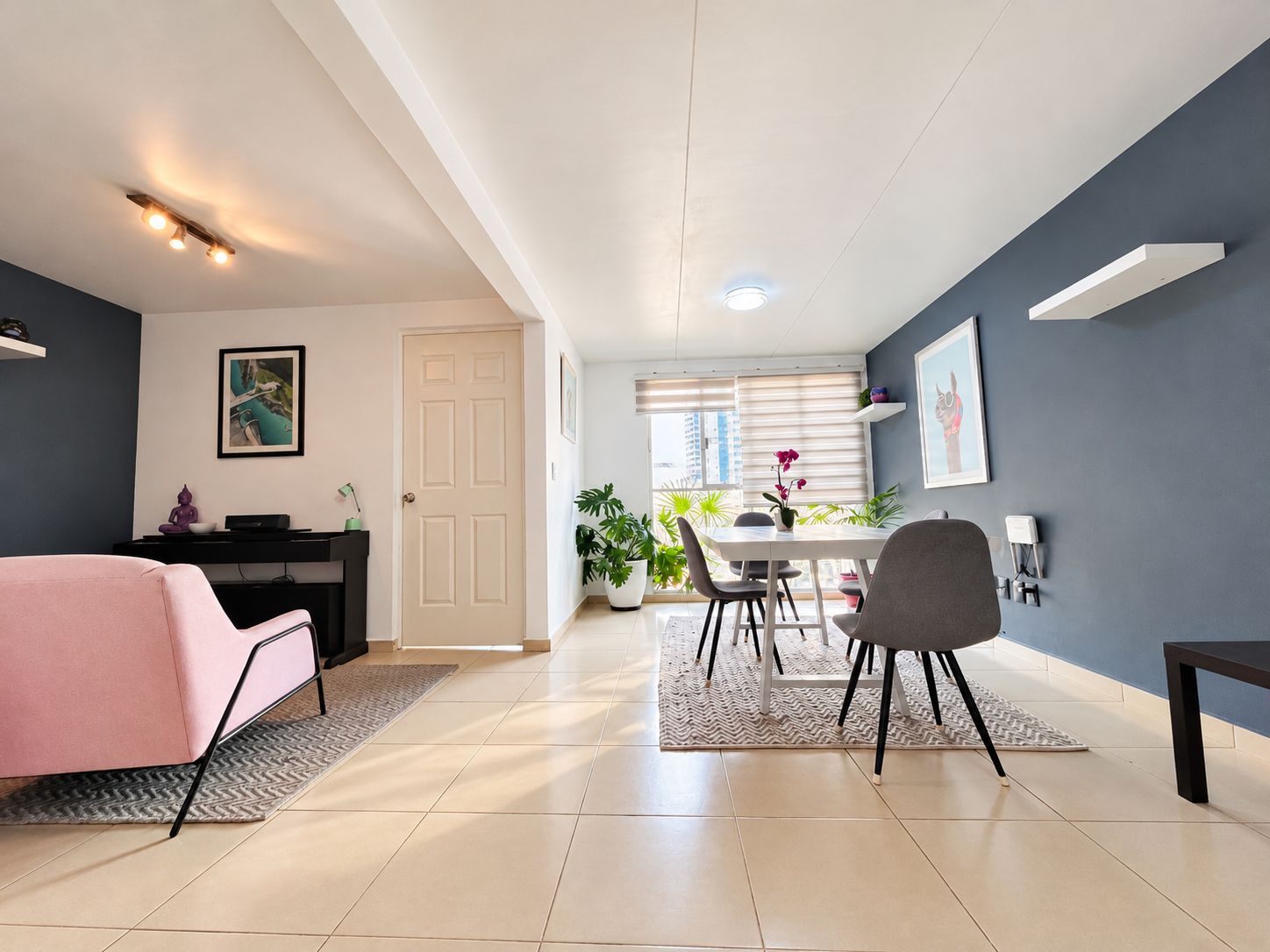 Un salon moderne avec un canapé, un coin repas comprenant une table et des chaises, un coin cuisine avec un réfrigérateur, et une porte donnant sur une autre pièce.