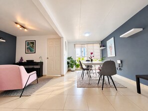 Una sala de estar moderna con un sofá, un comedor con mesa y sillas, una cocina con refrigerador, y una puerta que conduce a otra habitación.