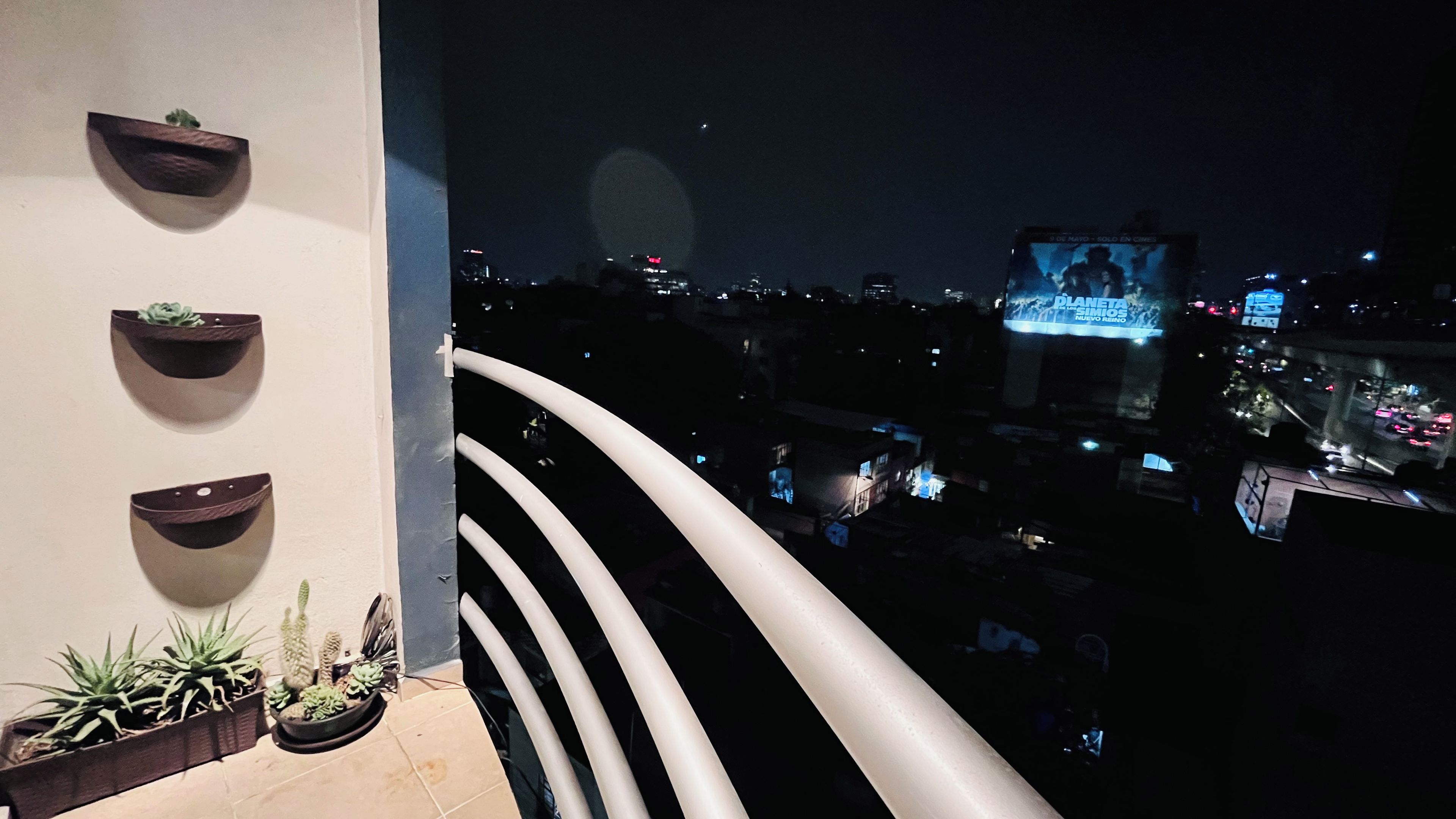 Een balkon met plantenbakken, een hekwerk en een uitzicht op de stad 's nachts.