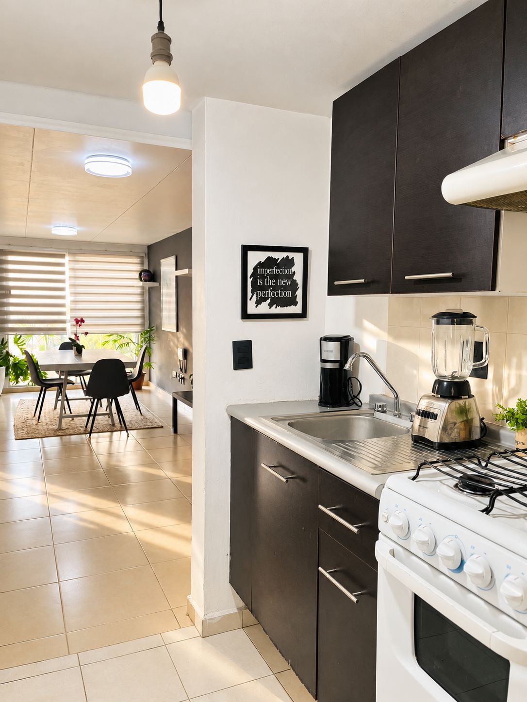 Une cuisine moderne avec des meubles de couleur sombre, une cuisinière, un mixeur et une machine à café. On y trouve également un coin repas avec une table et des chaises, ainsi qu’une fenêtre équipée de stores.