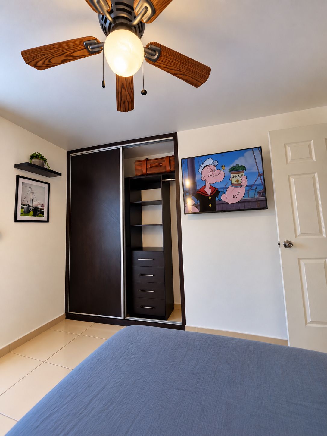 Une chambre à coucher avec un lit, un ventilateur de plafond, une armoire, une porte et un tableau encadré au mur.