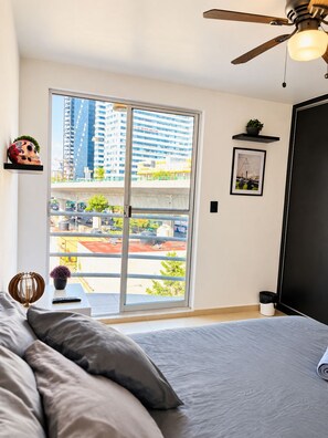 A bedroom with a bed, a ceiling fan, a window with a view of buildings, a shelf with a plant, and a framed picture.