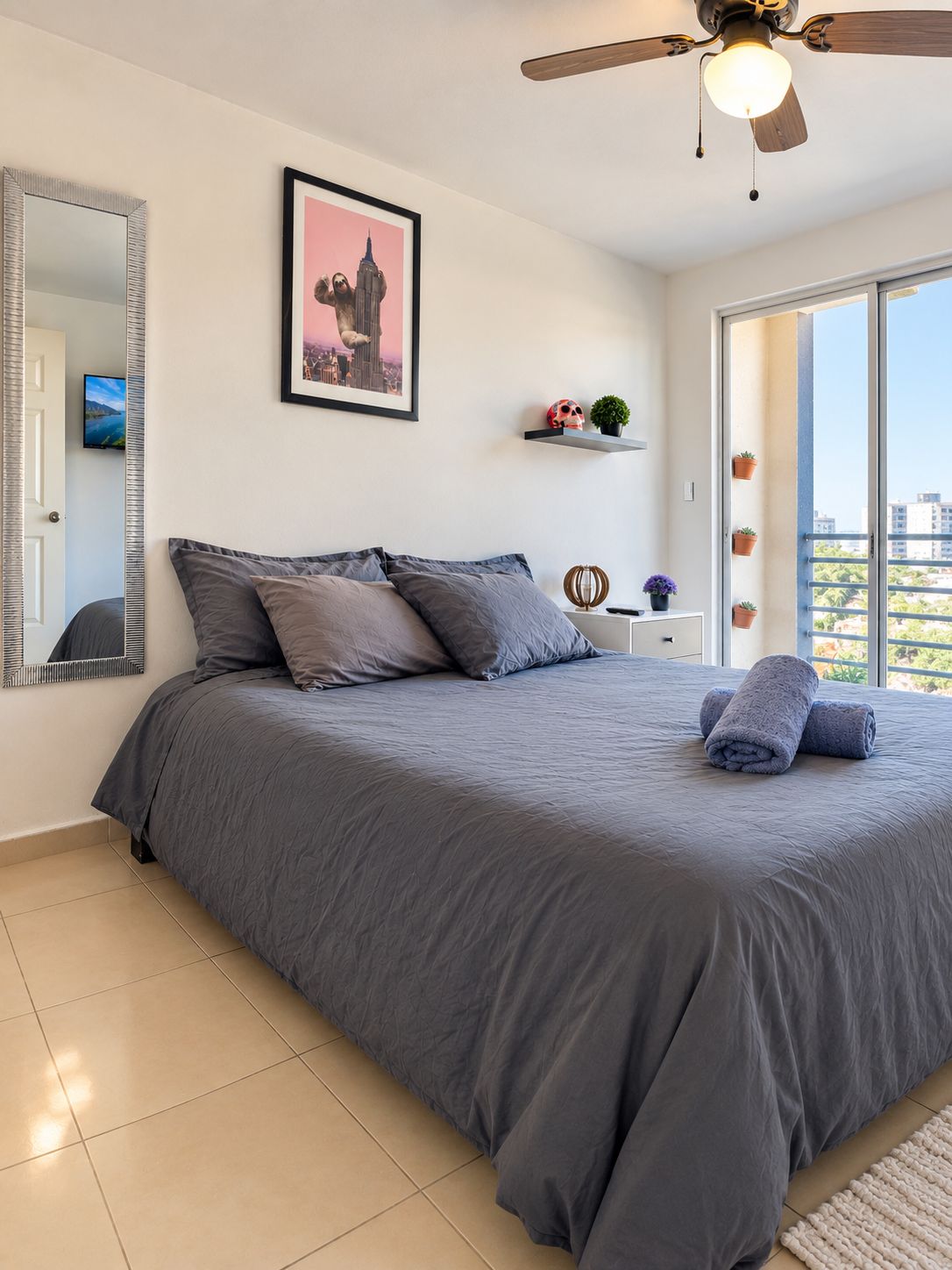 Une chambre avec un grand lit, un ventilateur de plafond, un tableau représentant un paysage urbain, une télévision et un balcon agrémenté de plantes en pot.
