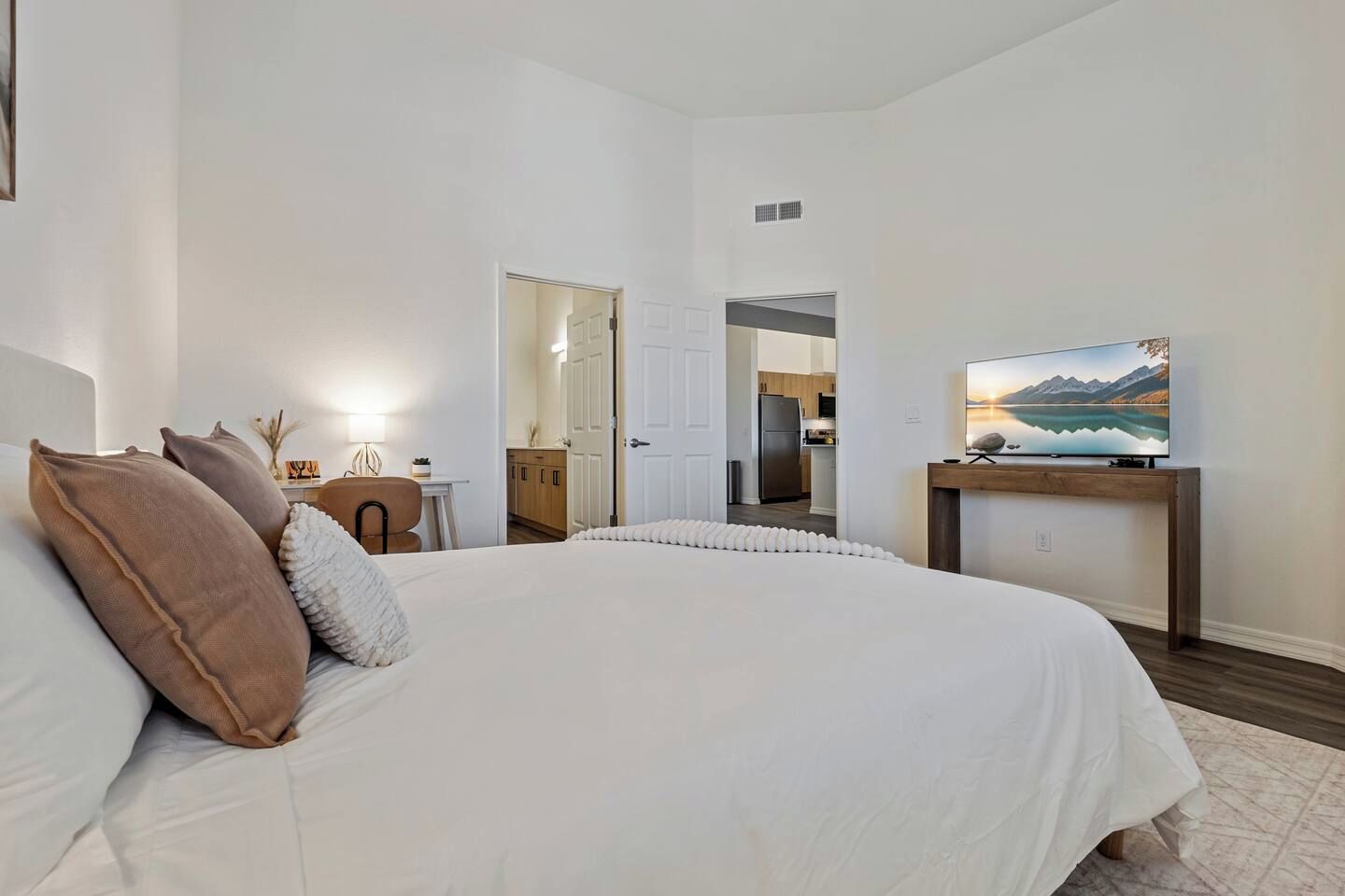 A bedroom with a large bed, a TV on a wooden stand, and a view into the kitchen.