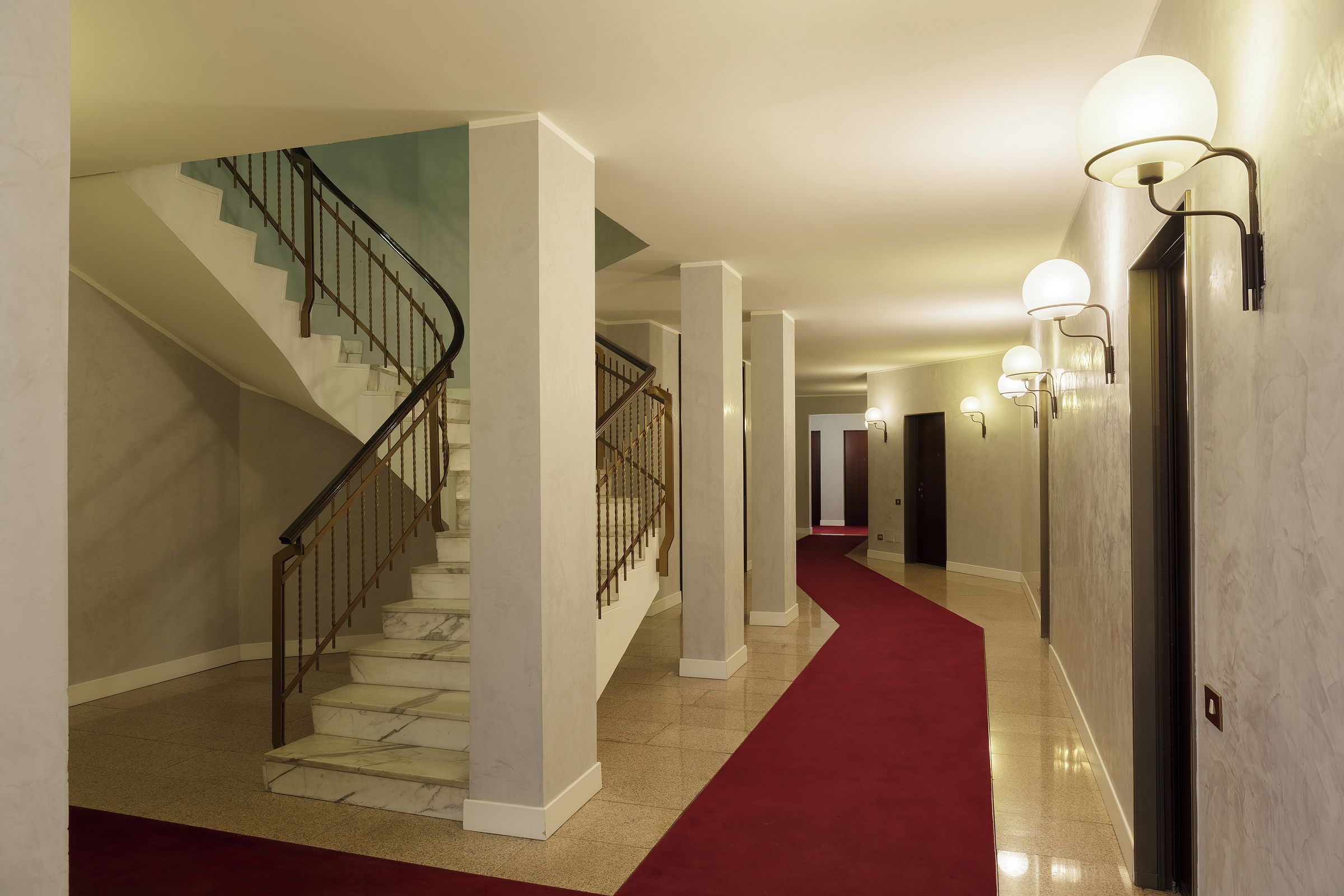 A hotel corridor with a marble staircase, white columns, and a red carpet.