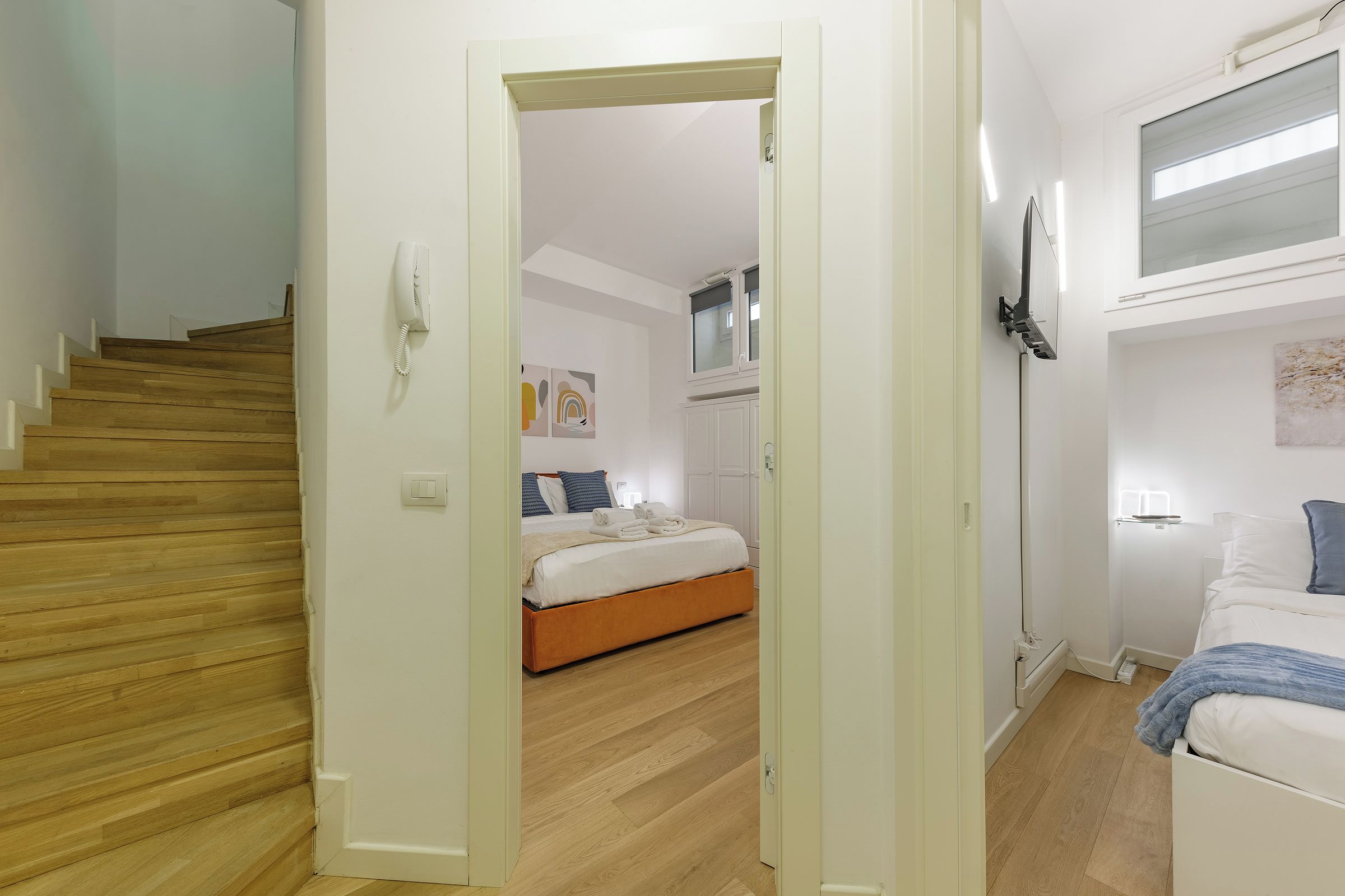A hotel room with a wooden staircase, a bed with white bedding, a bedside table, and a wall-mounted TV.