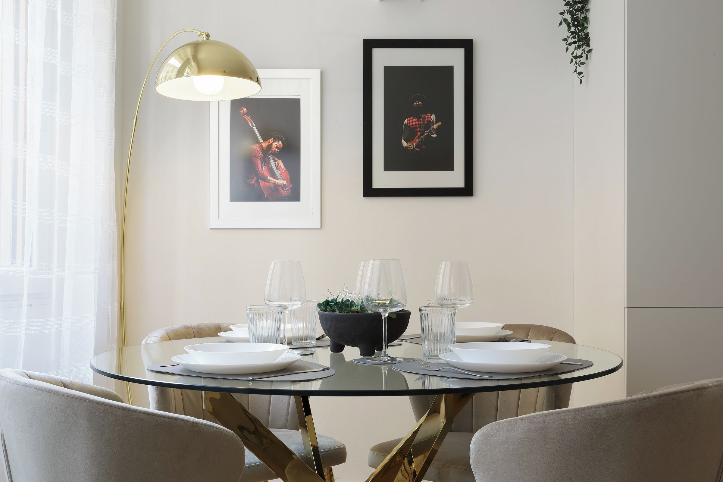 Un comedor con una mesa redonda de vidrio, cuatro sillas, un florero negro con plantas verdes y dos cuadros en la pared.