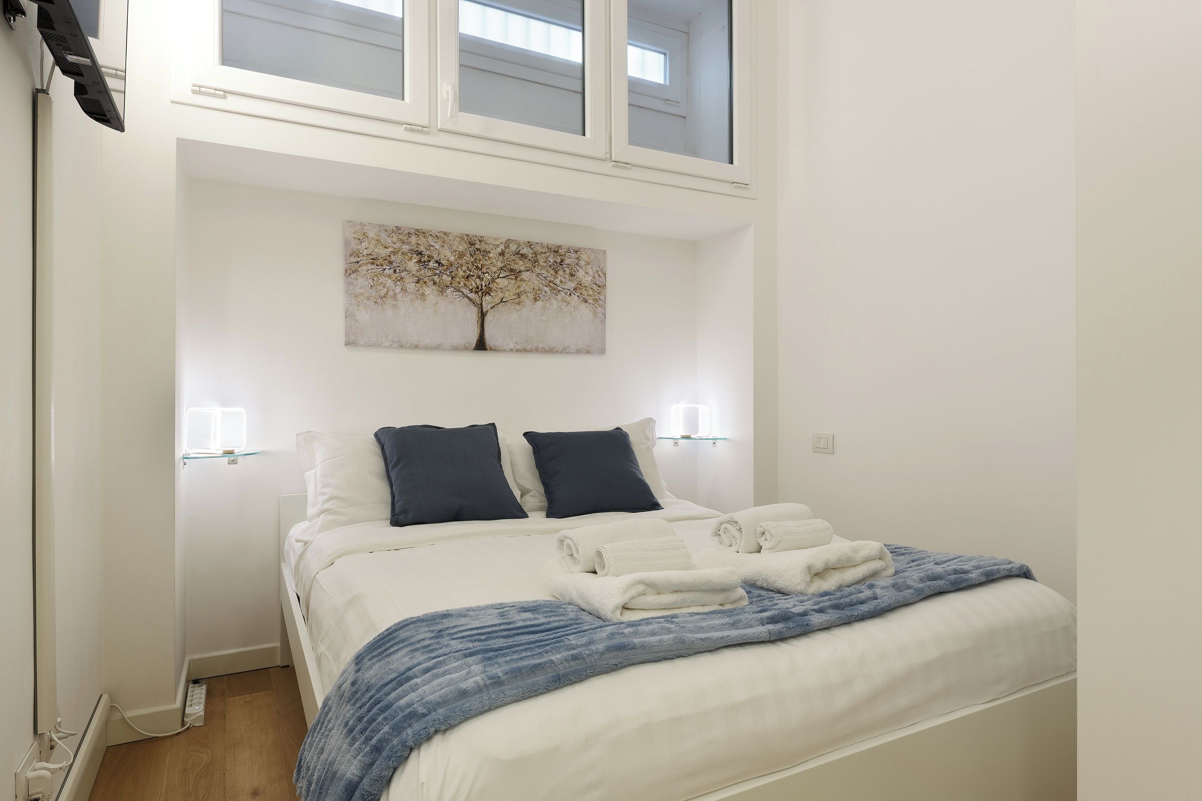 Une chambre à coucher avec un lit, deux lampes de chevet, un tableau représentant un arbre et une fenêtre située au-dessus du lit.