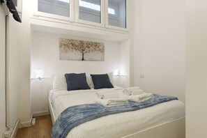Ein Schlafzimmer mit einem Bett, zwei Nachttischlampen, einem Baumgemälde und einem Fenster über dem Bett.