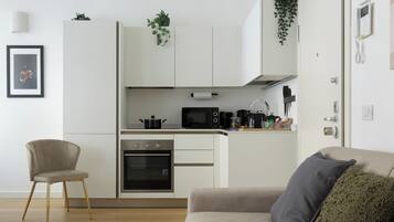 Una cocina moderna con armarios blancos, horno empotrado, microondas y cafetera. Hay un sofá color beige con un cojín verde y un cuadro en la pared.