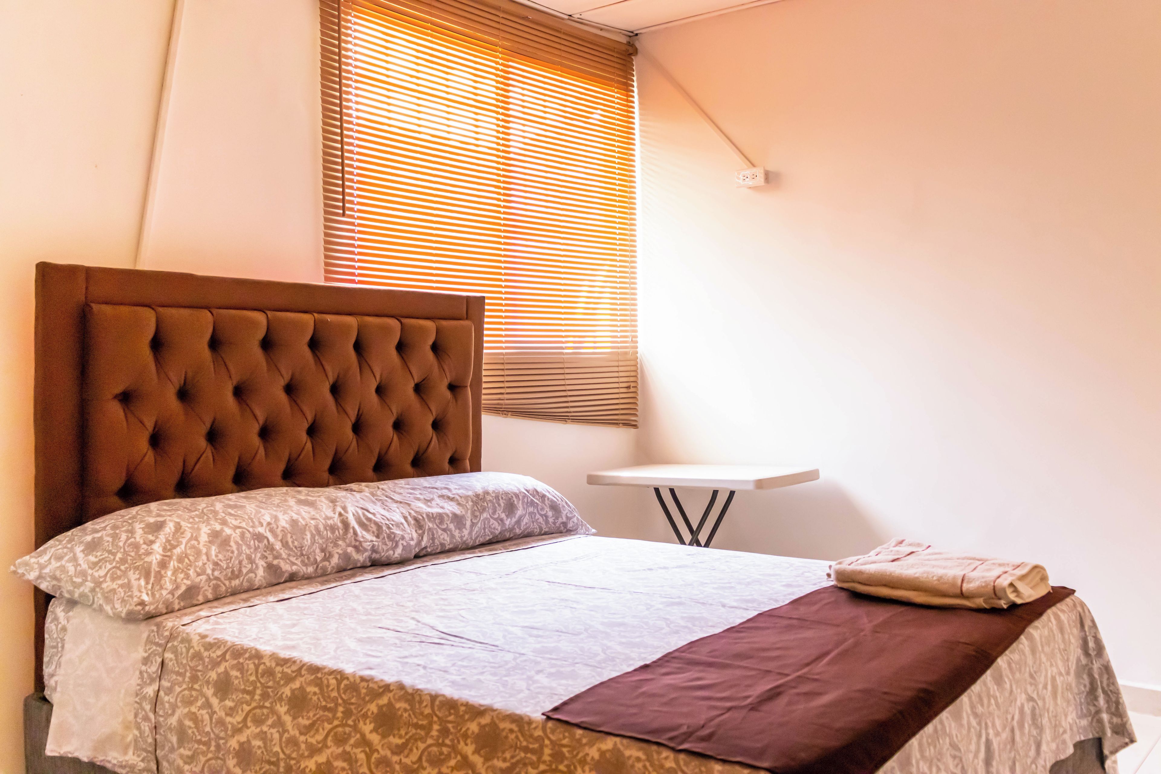 A neatly made bed with a tufted headboard, a bedside table, and a window with blinds.