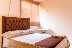 A neatly made bed with a tufted headboard, a bedside table, and a window with blinds. - Mirador del pastelillo (Bolívar)