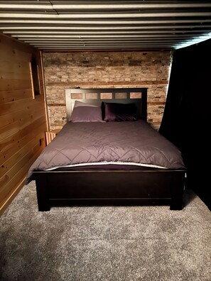Ein Einzelbett mit Kopfteil an einer Steinwand, eine Holzdielendecke und ein Teppichboden.