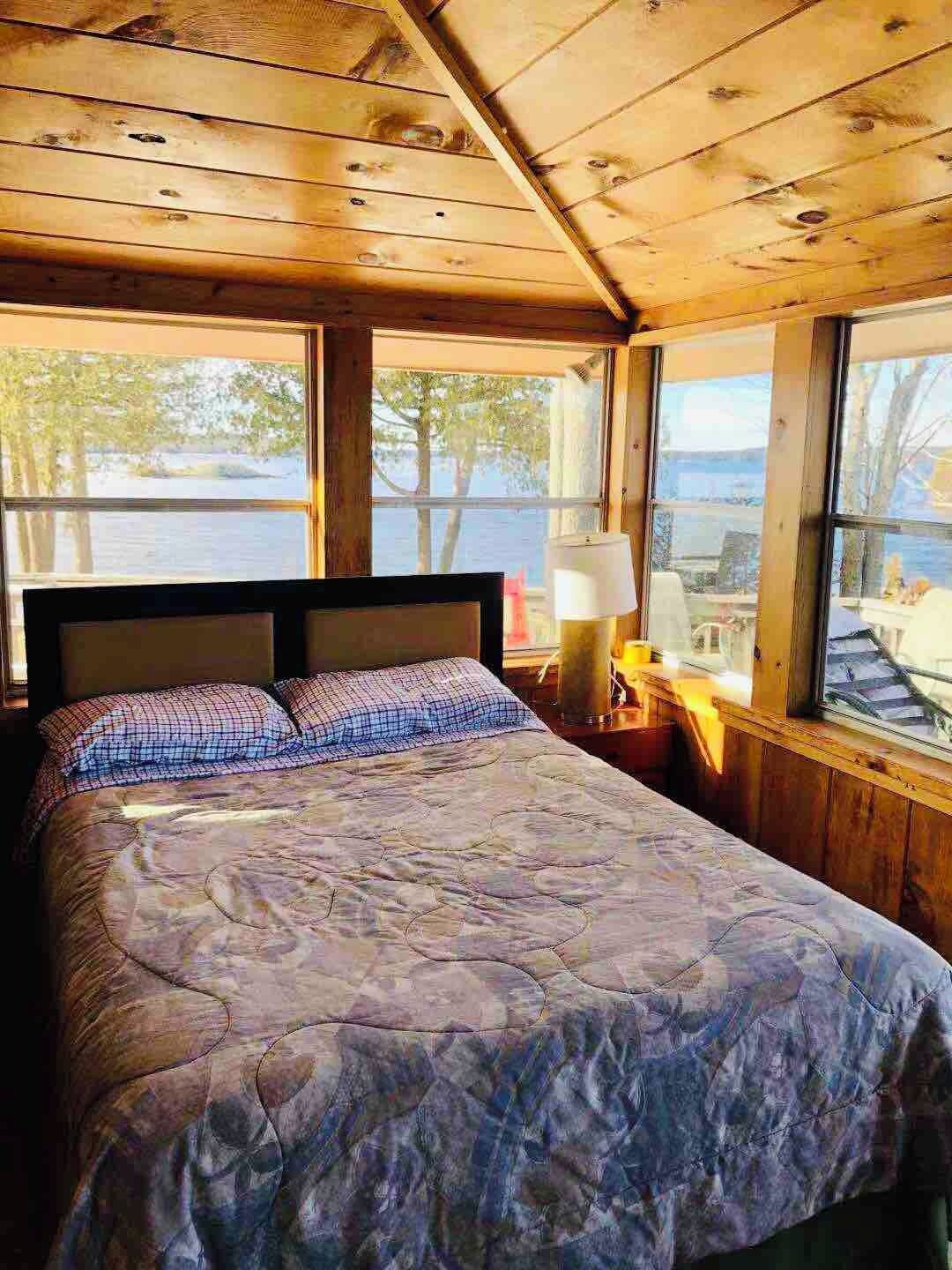 A wooden cabin room with a bed, a nightstand, and a window overlooking a body of water.