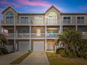 A multi-story residential building with a balcony on each floor, a garage at the front, and a well-maintained lawn. - The Bent Palm (Kure Beach)