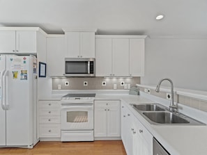 A modern kitchen with white cabinetry, a built-in microwave, a stainless steel sink, and a double oven. - The Bent Palm (Kure Beach)