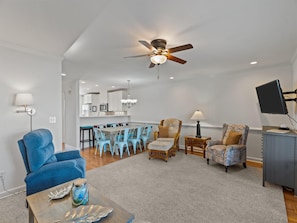 A modern living room with a dining area, a ceiling fan, and a kitchen in the background. - The Bent Palm (Kure Beach)