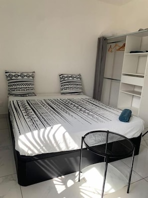 A bed with patterned bedding, a chair, a wardrobe, and a shelf unit in a room with white walls. - NGAYABE 976 (MAMOUDZOU)