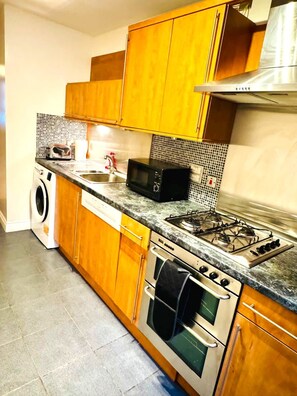 A kitchen with wooden cabinets, a microwave, a stove, and a washing machine. - Modern Finnieston 2 Bed Apt, near SEC & OVO, Free Parking (Glasgow)