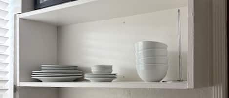 A kitchen with a white countertop, a sink, and a shelf above holding plates and bowls.