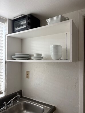 A kitchen with a white countertop, a sink, and a shelf above holding plates and bowls.