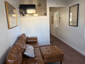 Un salon avec un canapé en cuir marron, un coussin blanc, un téléviseur à écran plat fixé au mur et, au fond, un coin cuisine.