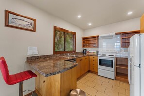 A kitchen with granite countertops, wooden cabinets, a stainless steel sink, a white stove, and a red chair. - Fern View - Pukawa Bay Holiday Home (Turangi)