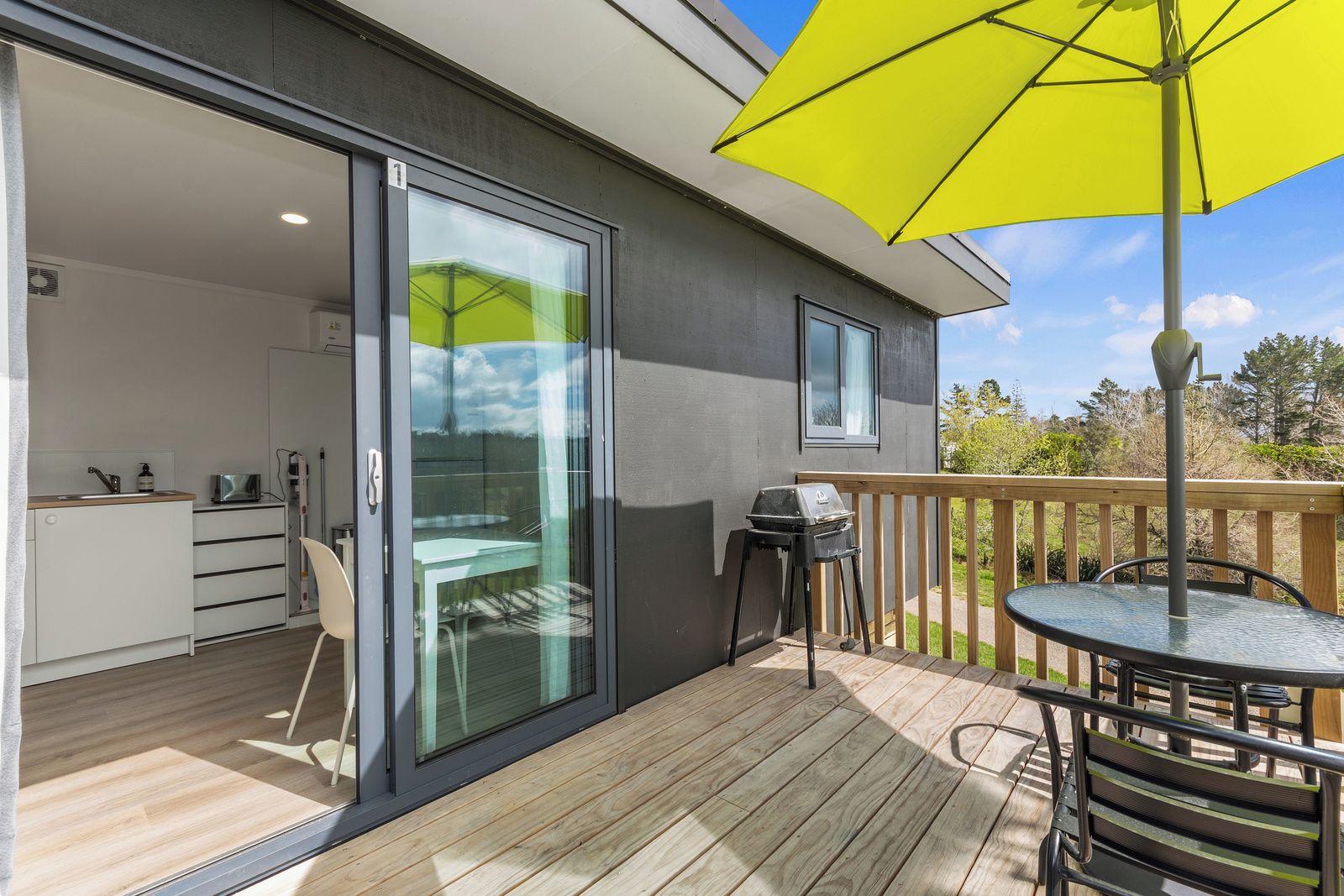 A modern outdoor deck with a sliding glass door leading to a kitchen area, a barbecue grill, and a table with a yellow umbrella.