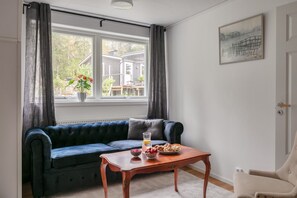 Ein Zimmer mit einem dunkelblauen Sofa, einem Holztisch mit Obstschale und einem Fenster mit Vorhängen.