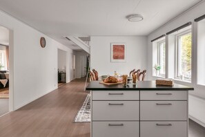 Eine moderne Küche mit einer zentralen Kochinsel, weißen Schränken und Blick ins Freie durch große Fenster.