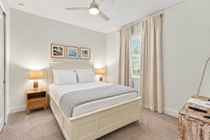 Quarto com cama, duas mesas de cabeceira, ventilador de teto, janela com cortinas e quadros nas paredes.
