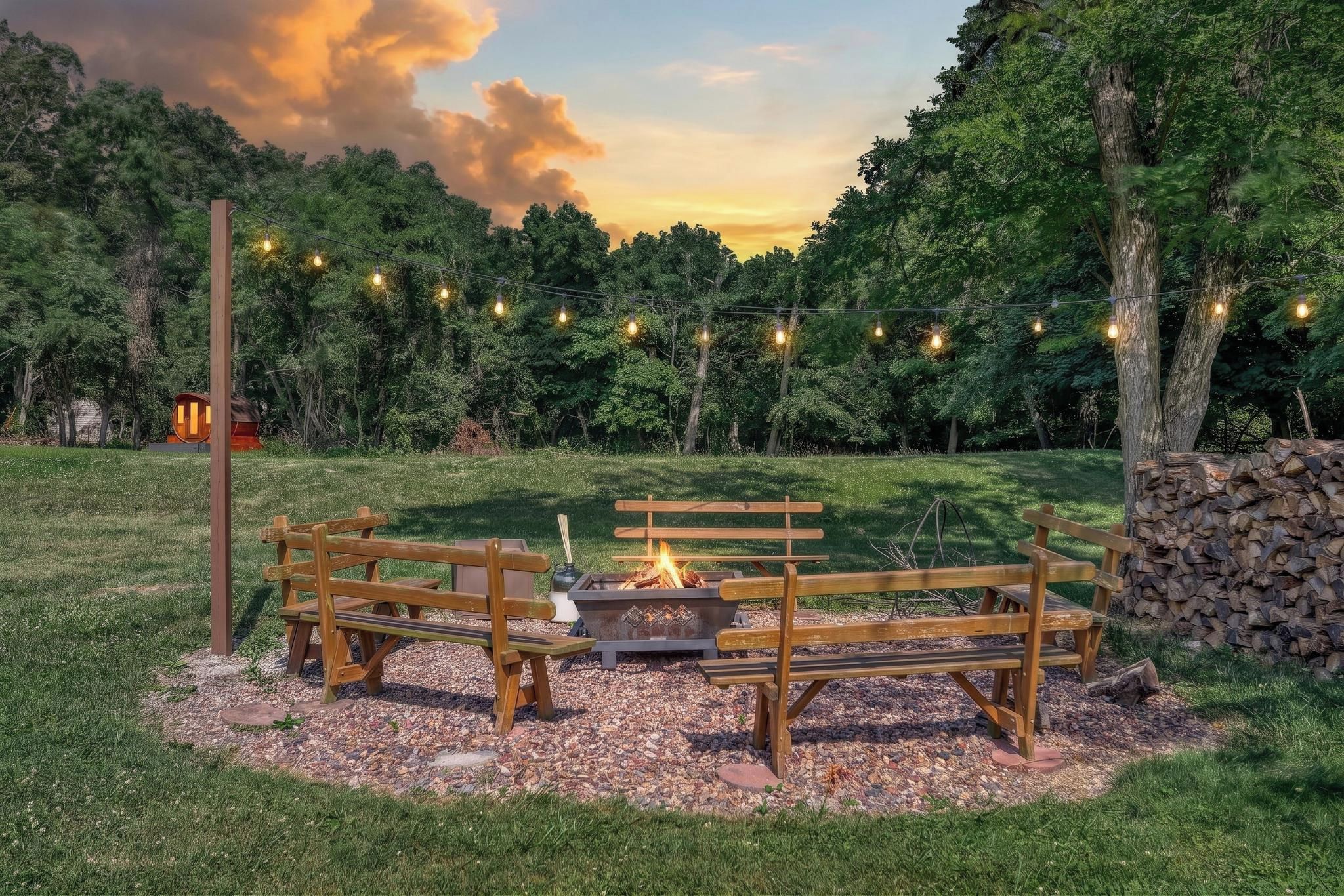 Un banc en bois et un foyer au milieu d’un espace extérieur verdoyant, agrémenté d’arbres et de guirlandes lumineuses.