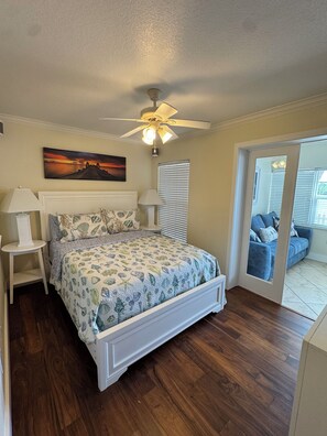 A bedroom with a bed, a ceiling fan, a nightstand, a lamp, a window with blinds, and a painting on the wall. - Waterfront Condo in Indian Shores- Beach - Pool - Boat Slips (Indian Shores)