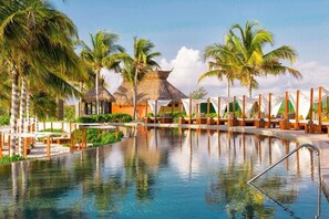 A pool area with lounge chairs and umbrellas, surrounded by palm trees and a thatched-roof building. - Two-Story Penthouse Loft, 3-bdrm Beachfront at Villa del Palmar Luxury Resort (Cancun)