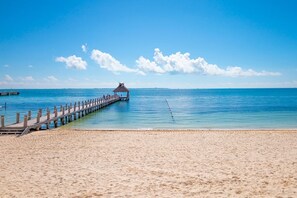 Ein Strand mit einer Holzstege, der zu einem Gebäude mit einem Reetdach führt.