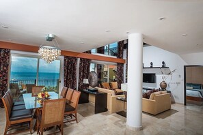 A modern living room with a dining area, a large window with a view, and a flat-screen TV. - Two-Story Penthouse Loft, 3-bdrm Beachfront at Villa del Palmar Luxury Resort (Cancun)
