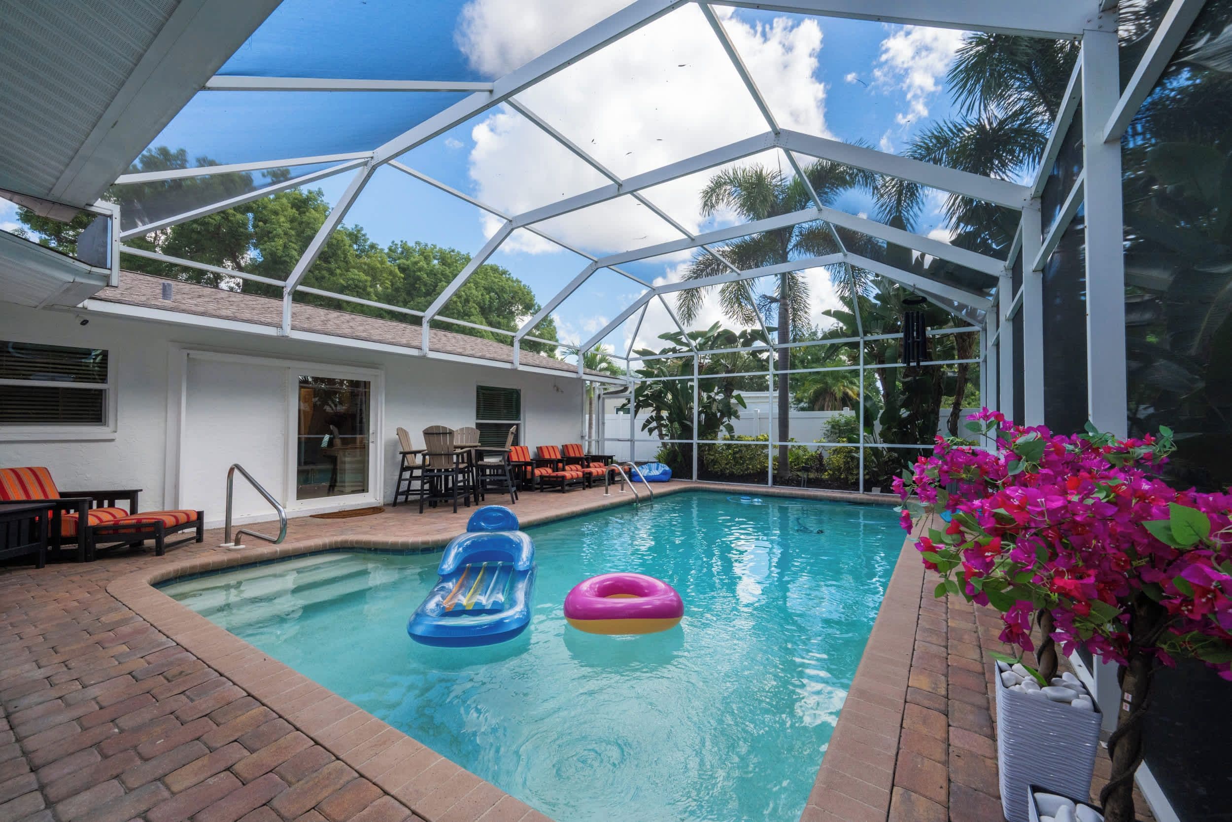 An indoor swimming pool with clear blue water, surrounded by a white roof structure, and furnished with lounge chairs and a small table.