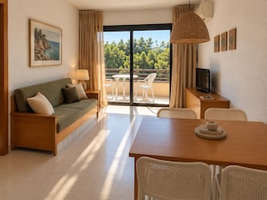 A room with a sofa, a dining table, chairs, a TV, and a balcony with a view of trees. - Golf Beach Apartment With Pool for 2 (Pals)