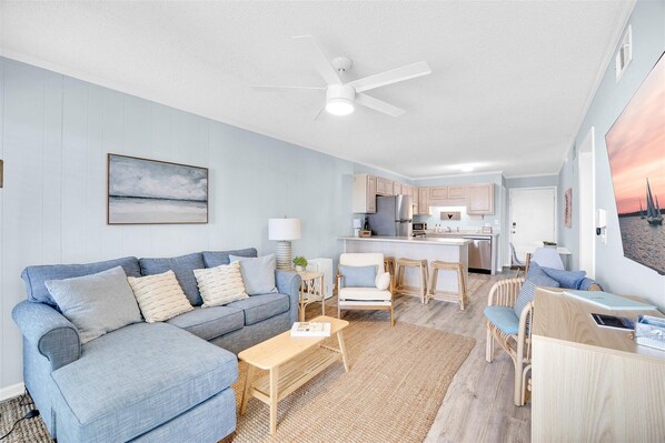 Ein modernes Wohnzimmer mit einer Couch, einem Couchtisch und Sesseln, eine Küche mit Kochinsel und Deckenventilator.