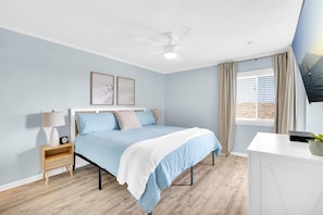 Un dormitorio con cama, mesitas de noche, ventilador de techo, ventana con cortinas y televisor en la pared.