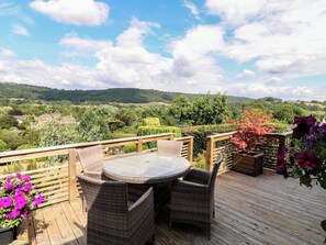 A wooden deck with a round table and wicker chairs, overlooking a lush landscape with hills and trees. - Hillset View (Bakewell)